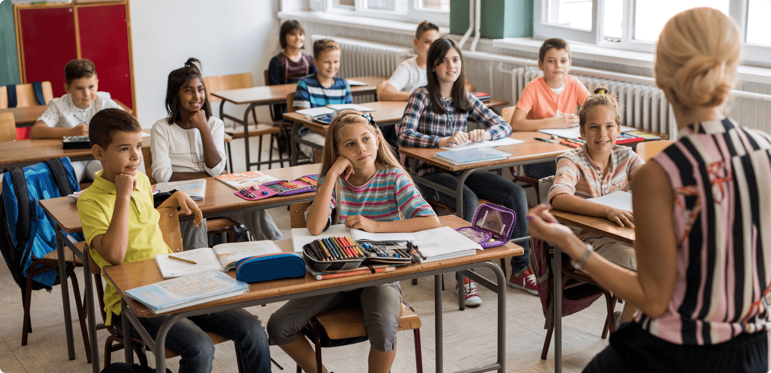 a group of young students sitting at desks in a classroom, looking intently at a blonde-haired female teacher.