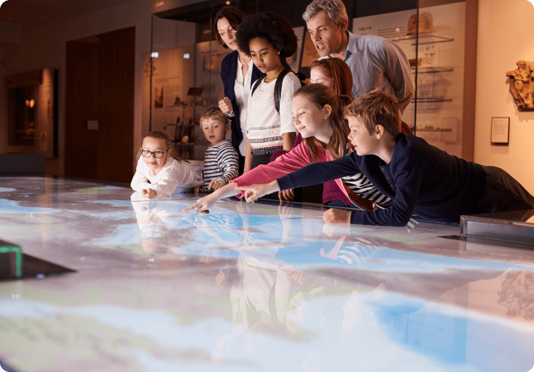 a mixed aged group of children and teens at a museum. Two adult teachers stand above them. The children are pointing at a touchscreen table
