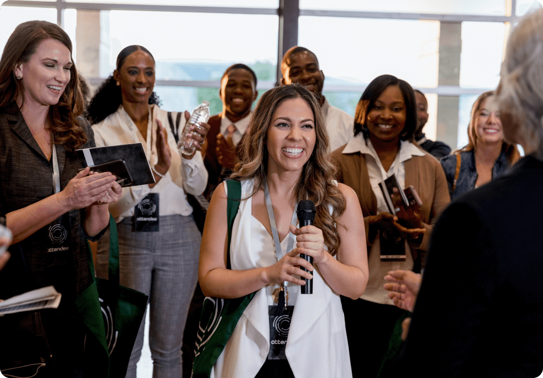 A group of adults at a conference applauding as someone is given recognition. There is a woman with wavy brown hair in the middle smiling and holding a microphone