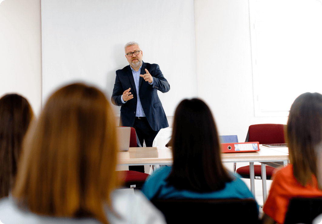 an older white man wearing glasses in the middle of a presentation to a group of teenaged students.