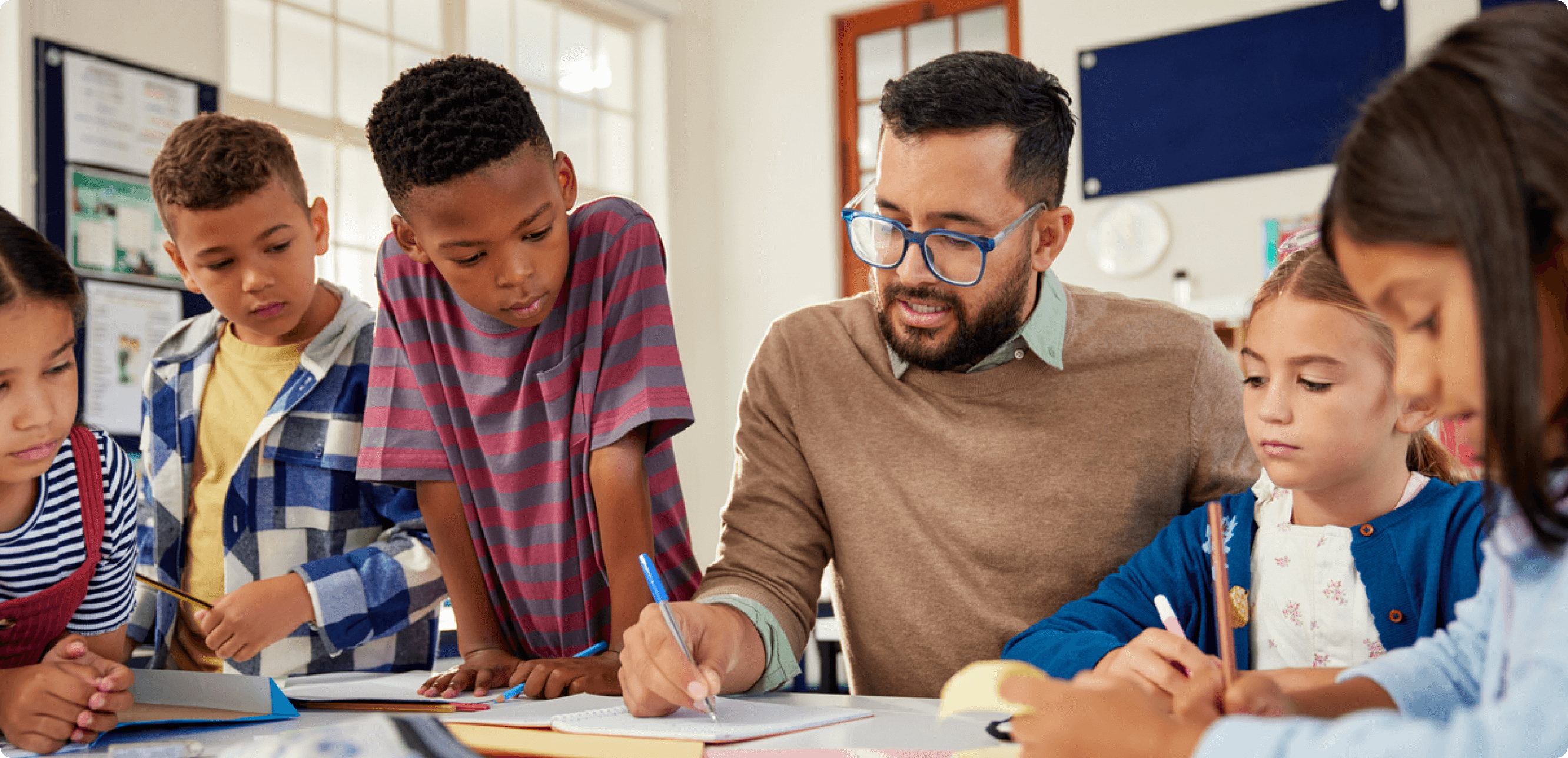 a bearded male teacher writing in a notebook with a pen, surrounded by young school children watching him intently