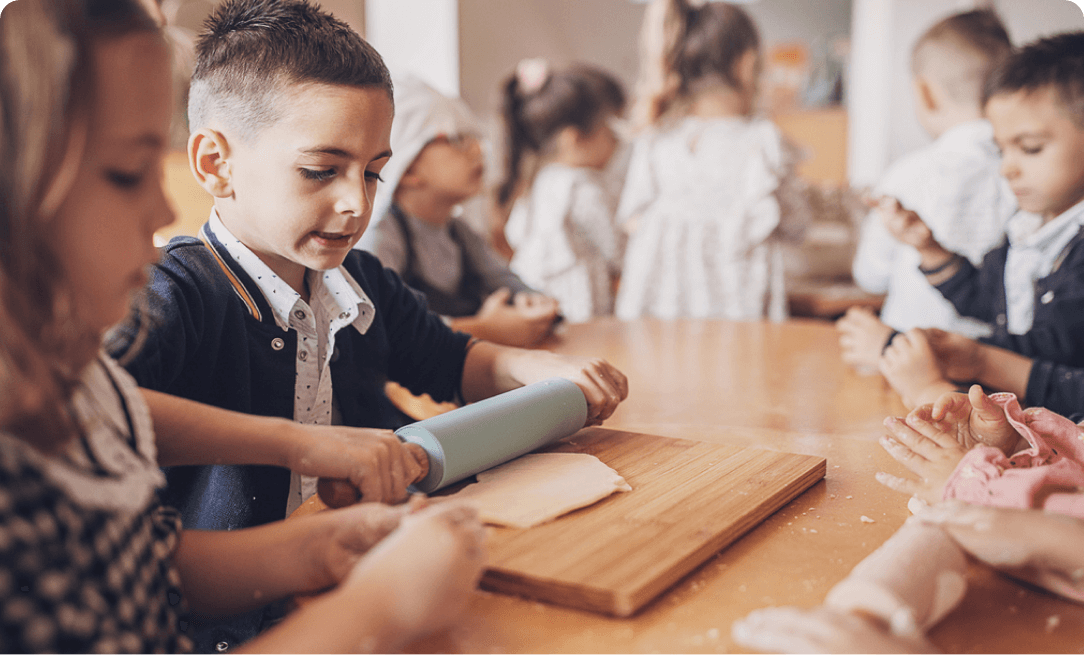 a young boy rolling pastry dough with a rolling pin. He is surrounded by similarly aged children also working with dough