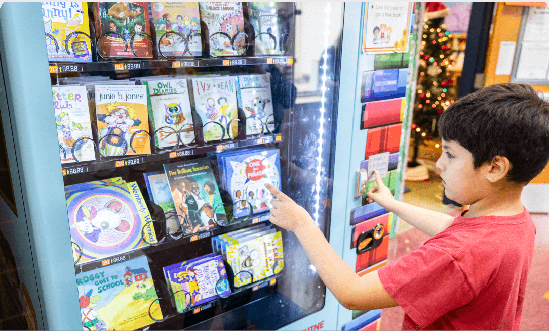 a young boy wearing a red shirt pointing to a selection at a book vending machine