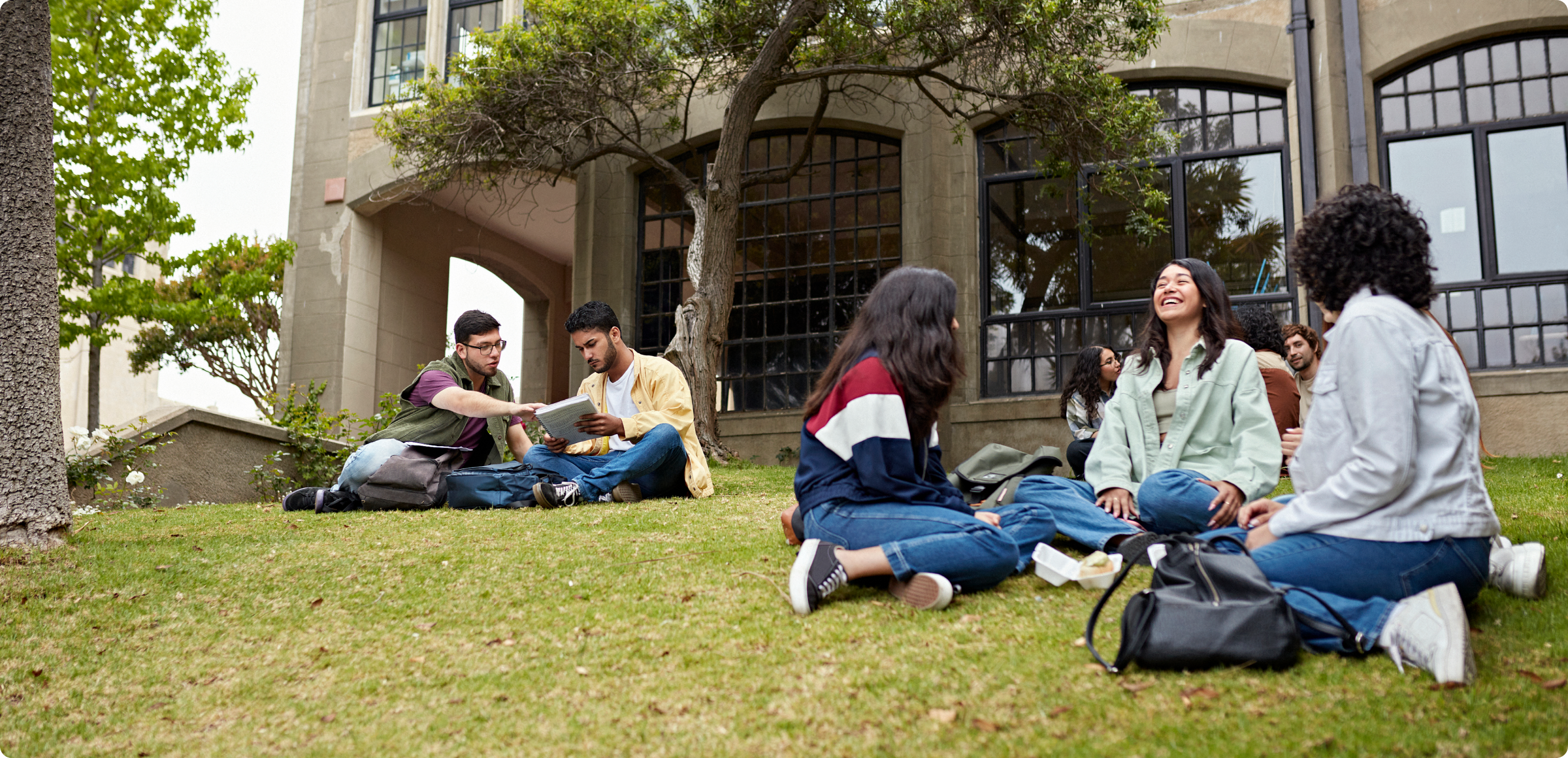 a group of older teens or young adults sitting outside on the grass, bags around them, in conversation and looking at a textbook
