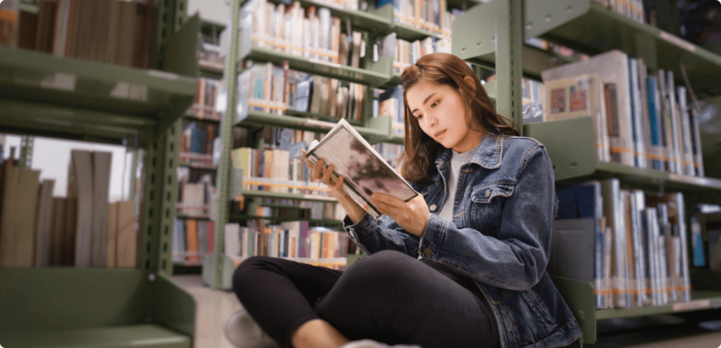 an older teenage girl sitting on the floor of a library, intently reading a book