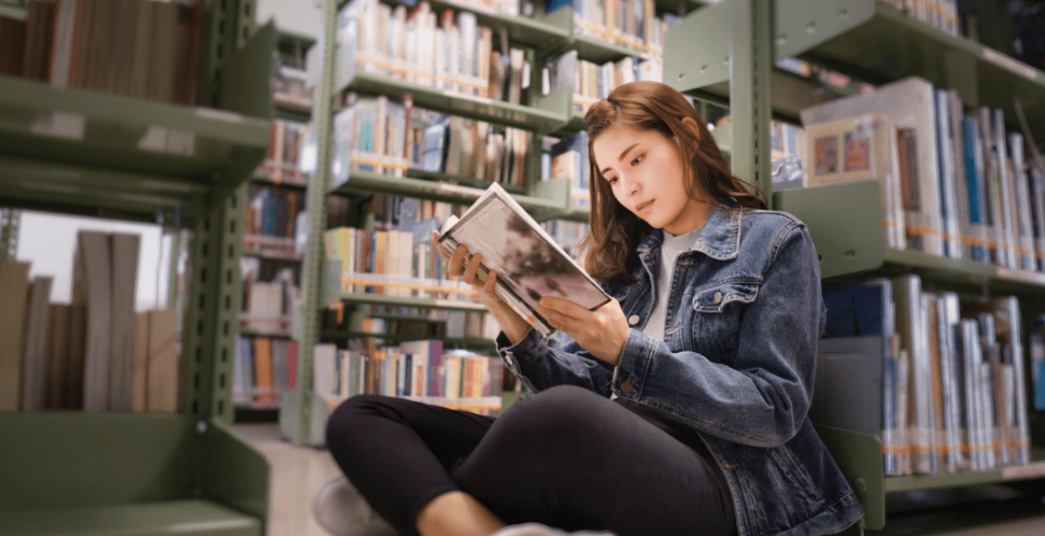 an older teenage girl sitting on the floor of a library, intently reading a book