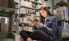 an older teenage girl sitting on the floor of a library, intently reading a book