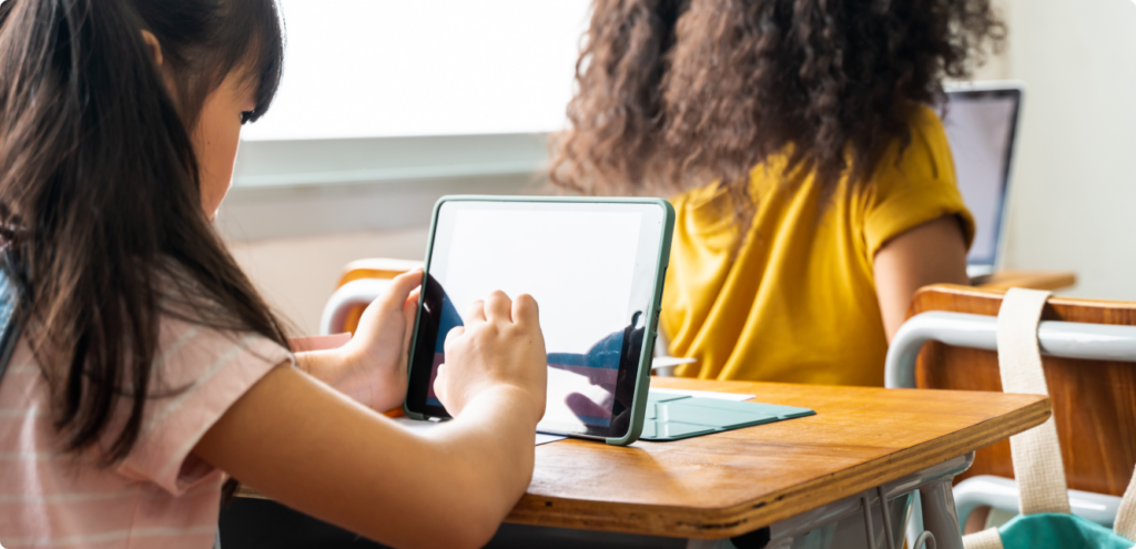 the back of a girl's head sitting at a school desk. On the desk sits an upright tablet, which the girl is tapping