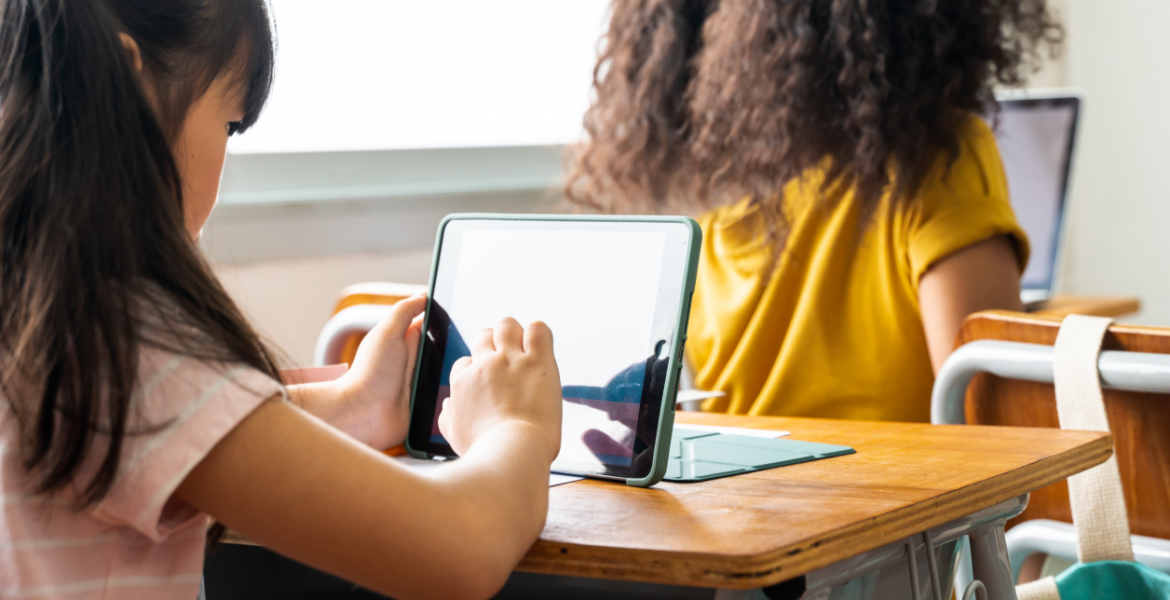 the back of a girl's head sitting at a school desk. On the desk sits an upright tablet, which the girl is tapping