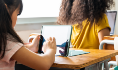 the back of a girl's head sitting at a school desk. On the desk sits an upright tablet, which the girl is tapping
