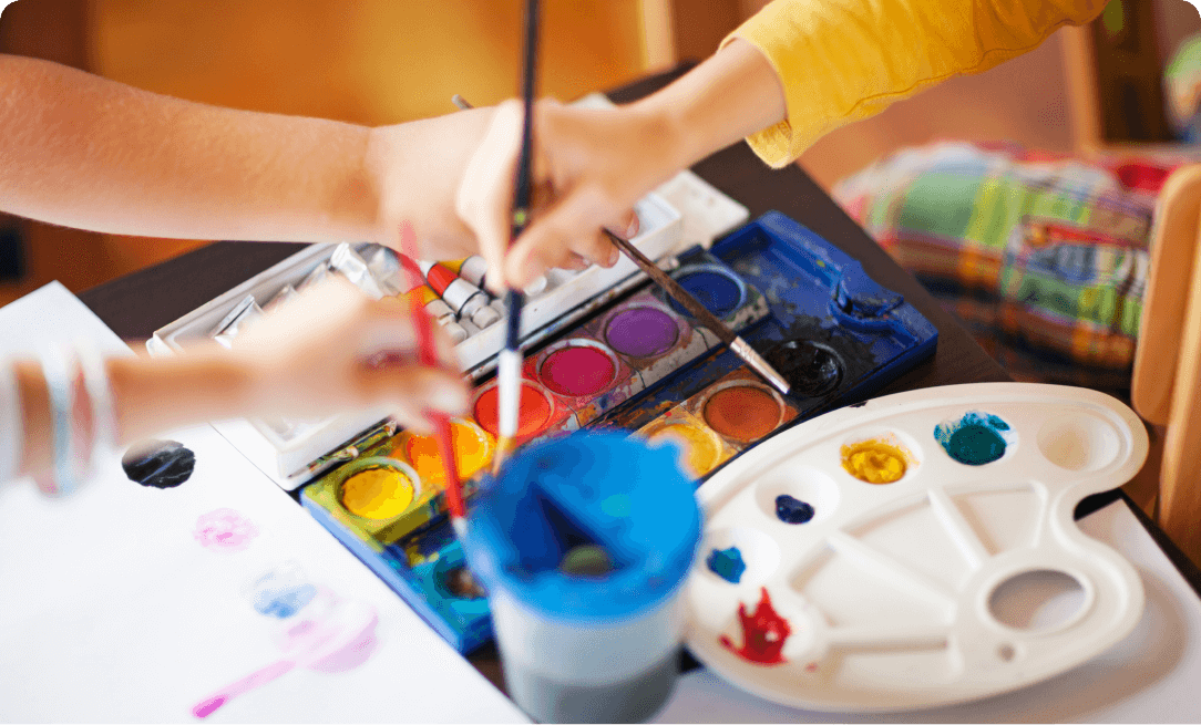 close up of hands of children holding paintbrushes dipping into a tray of paints