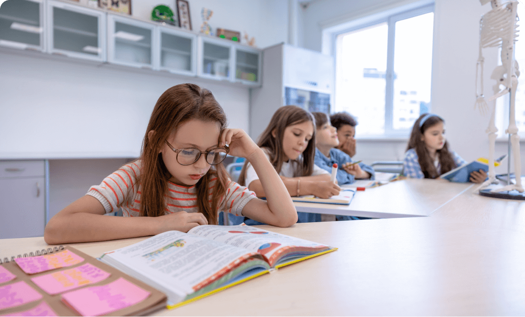 a young girl wearing glasses sitting at a desk, studying a school workbook. Several other young students are behind her, also studying.
