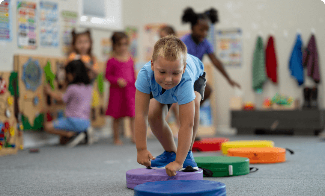 a young boy wearing blue at the center of the image, several young girls in the background out of focus. The boy is jumping on a series of multi-colored circular pads.