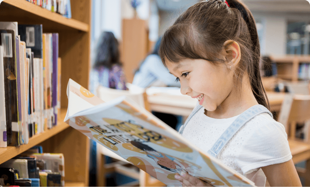 close up of a young Hispanic girl reading a picture book. The girl is wearing overalls and smiling