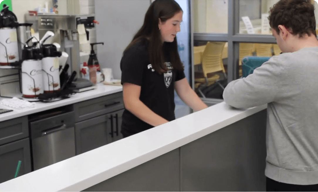 a teenage girl behind a coffee shop counter, wearing a black shirt with a logo on it, taking the order of a teenage boy