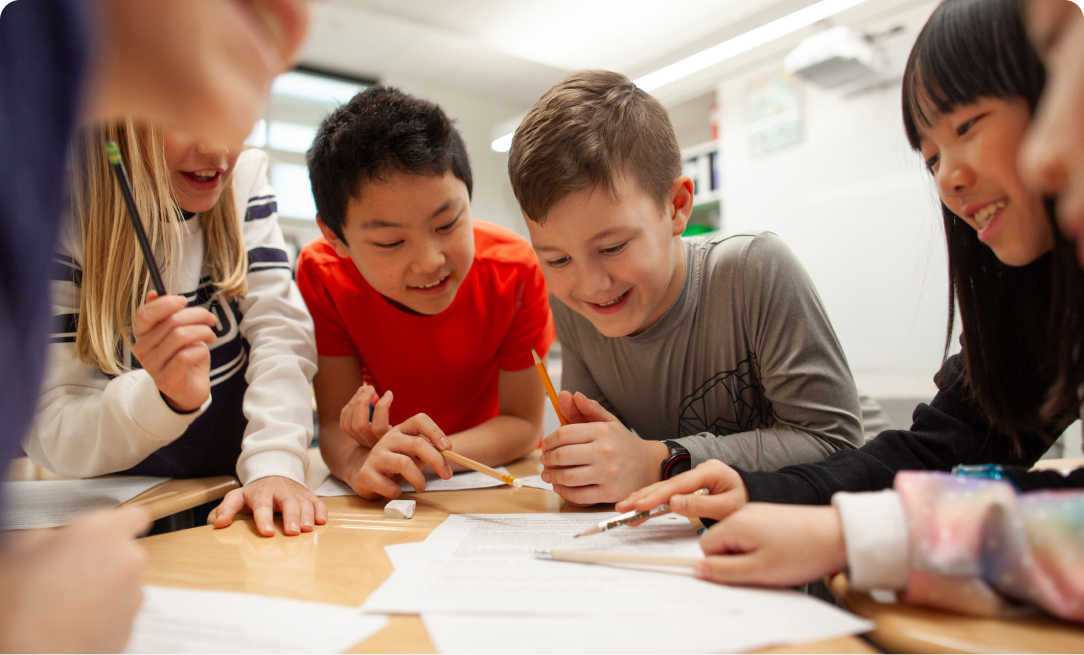 a group of children, mixed genders, sitting at a table circling around a worksheet. The children are holding pencils and pointing at the sheet of paper