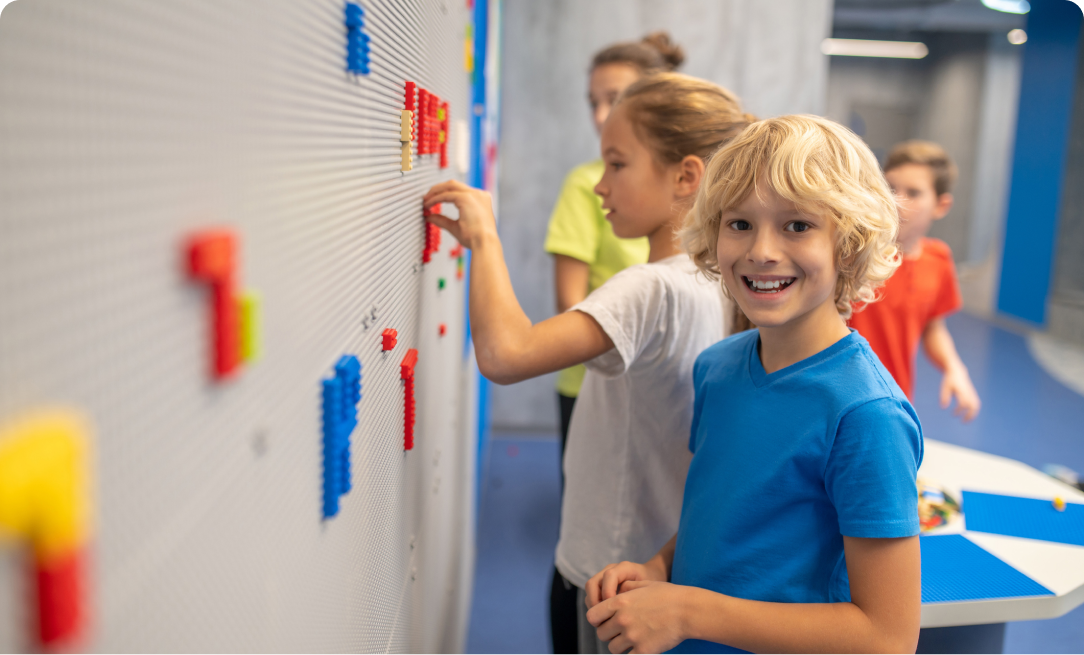 a young blonde boy wearing a blue shirt standing against a wall, letters are on the wall made of LEGO