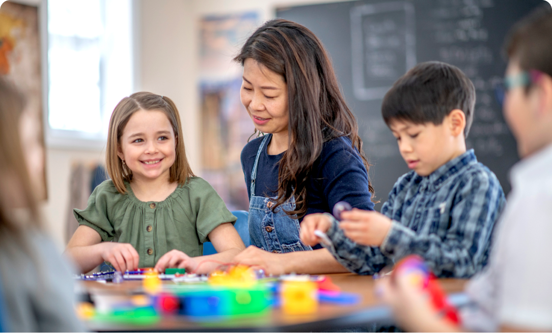 a female teacher sitting at a table helping female and male young students. The students are playing with blocks on the table