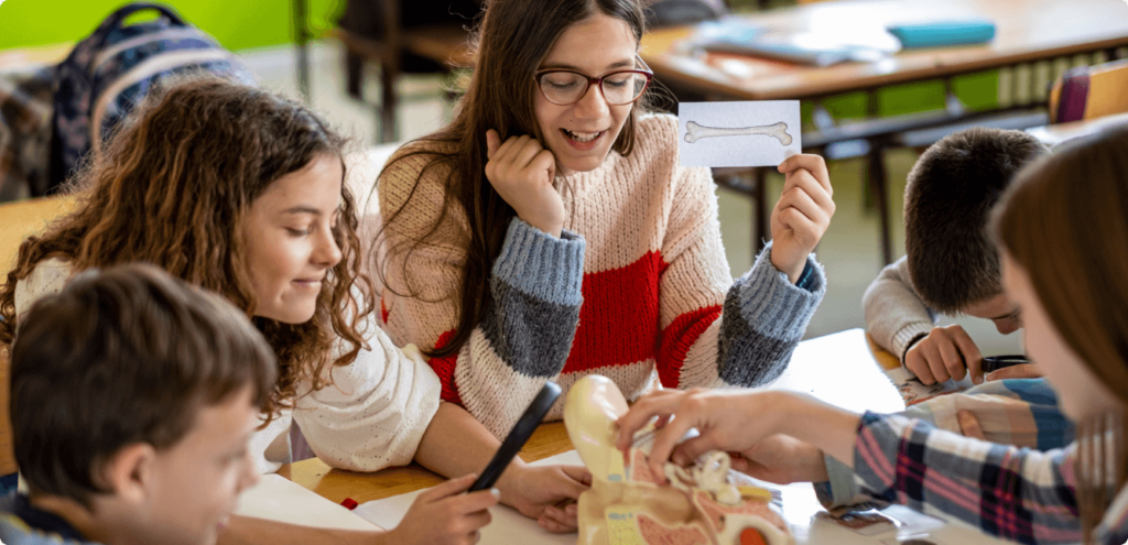 a group of teenagers around an anatomical model. A brown-haired teenage girl wearing glasses holds up a card with a drawing of a bone on it. Other teenagers examine the model in a hands-on manner