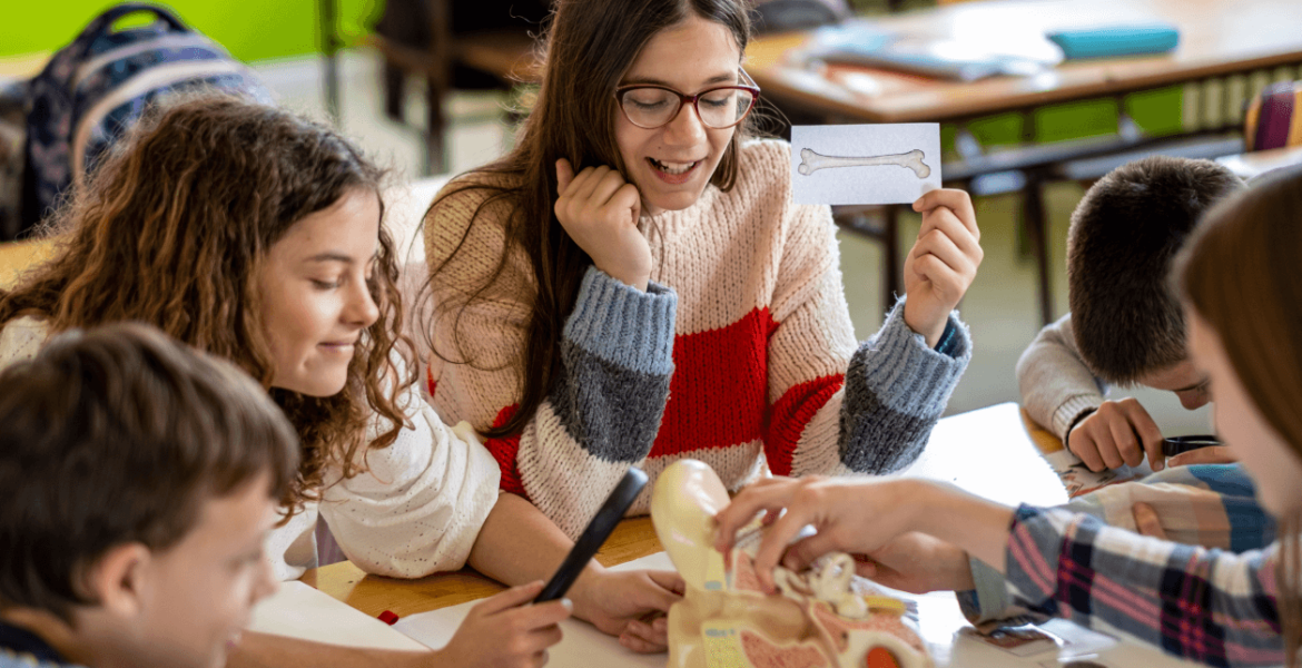 a group of teenagers around an anatomical model. A brown-haired teenage girl wearing glasses holds up a card with a drawing of a bone on it. Other teenagers examine the model in a hands-on manner