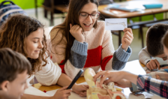 a group of teenagers around an anatomical model. A brown-haired teenage girl wearing glasses holds up a card with a drawing of a bone on it. Other teenagers examine the model in a hands-on manner