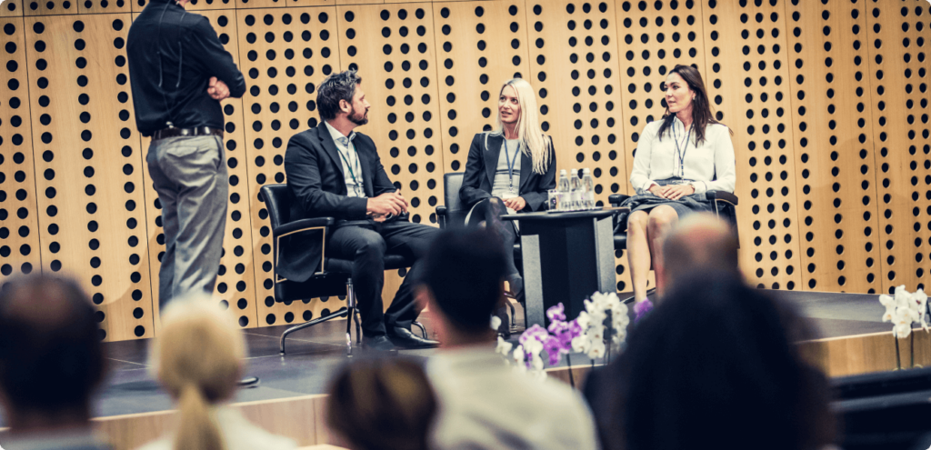 two adult women and one man sit on top of a stage at a conference. A discussion is currently being had between the three of them
