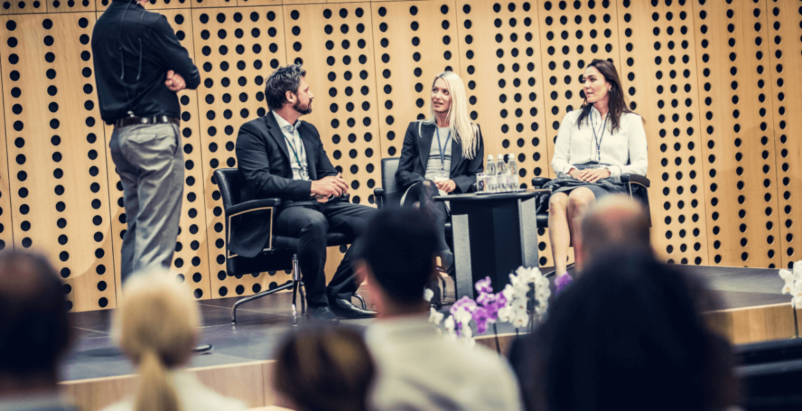 two adult women and one man sit on top of a stage at a conference. A discussion is currently being had between the three of them
