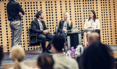 two adult women and one man sit on top of a stage at a conference. A discussion is currently being had between the three of them