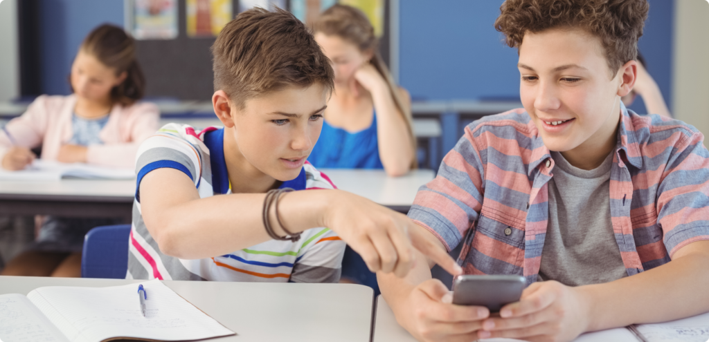 two teenage boys sitting at a school table with an open notebook in front of them. The boys are looking at a phone for information