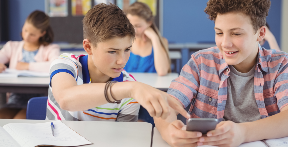 two teenage boys sitting at a school table with an open notebook in front of them. The boys are looking at a phone for information