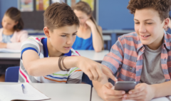 two teenage boys sitting at a school table with an open notebook in front of them. The boys are looking at a phone for information
