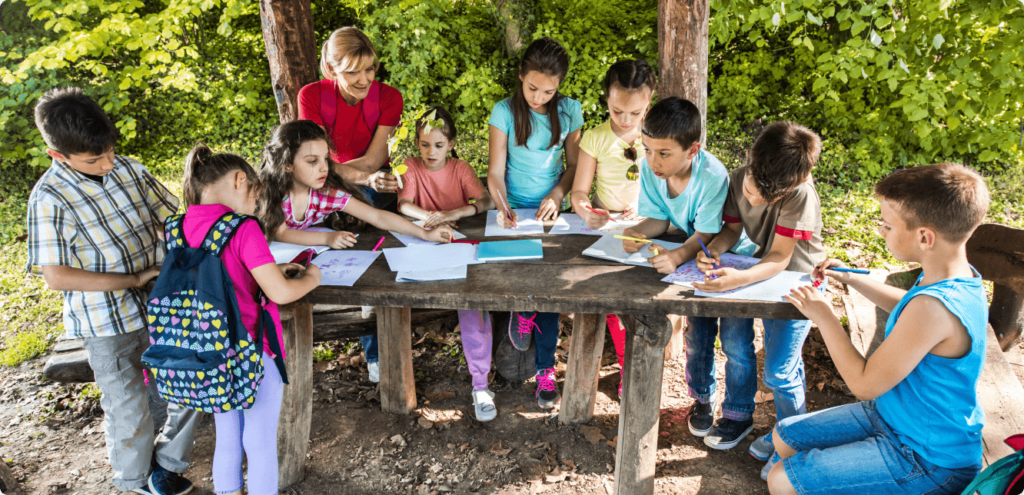 a group of young students and their teacher working outside at a wooden table. The children appear to be coloring with markers and pen