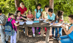 a group of young students and their teacher working outside at a wooden table. The children appear to be coloring with markers and pen